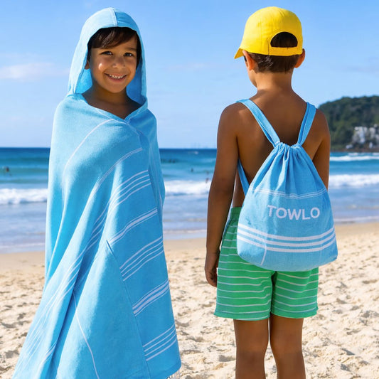 Boy wearing a Towlo kids hooded beach towel and drawstring backpack in aqua blue with white stripes at the beach, showing towel worn as poncho and backpack with Towlo logo