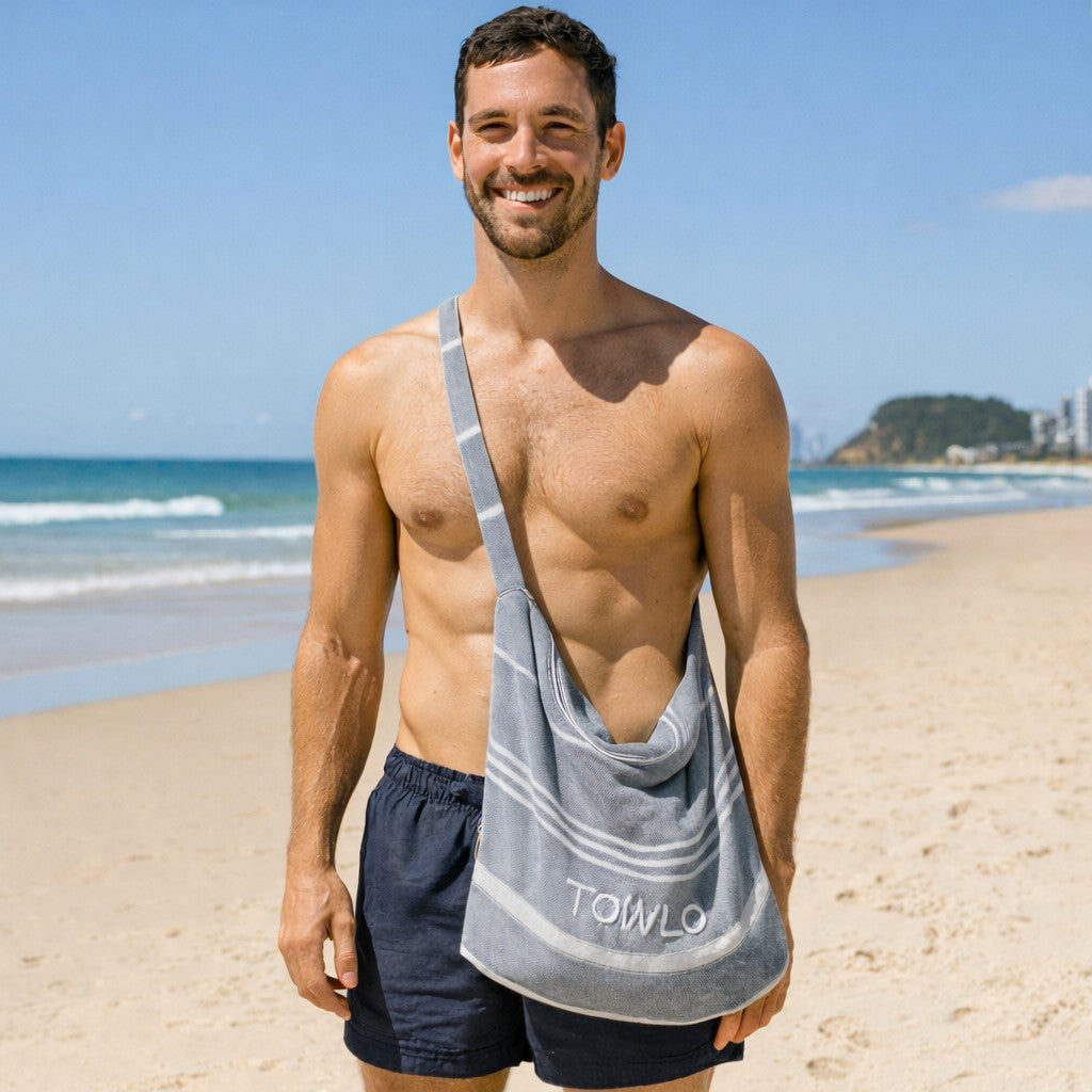Smiling man at the beach wearing a Towlo convertible towel tote bag crossbody, a lightweight beach towel that folds into a shoulder bag