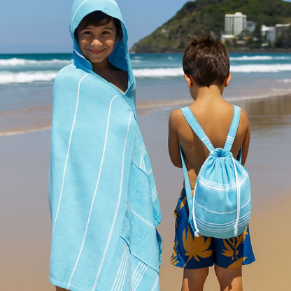 Two children at the beach wearing Towlo blue kids hooded towel backpacks, showcasing built-in hood, built-in towel, lightweight quick-dry fabric, and backpack design.
