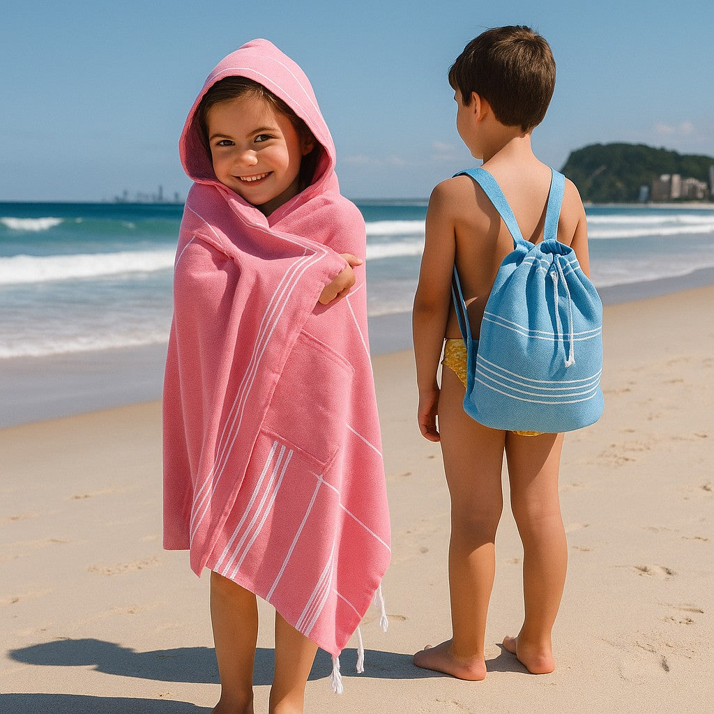 Young girl wrapped in a pink Towlo hooded towel standing on the beach beside a boy wearing a blue Towlo kids’ backpack towel, with ocean waves in the background.