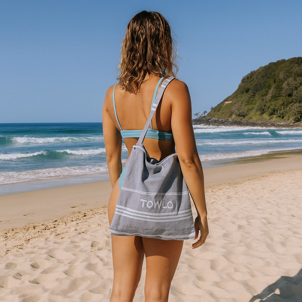 Woman walking on the beach wearing a Towlo convertible towel tote bag, a lightweight beach towel that folds into a shoulder bag