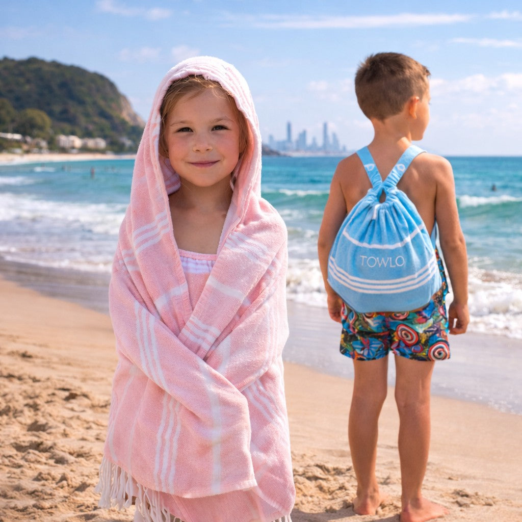 Two children on a beach, one wrapped in a pink towel and the other with a blue backpack, with a city skyline in the background.