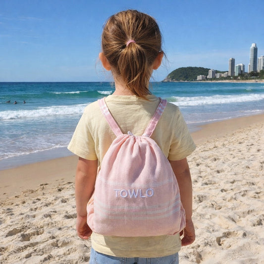 Child with a pink backpack labeled 'TOWLO' on a beach with ocean and city skyline in the background