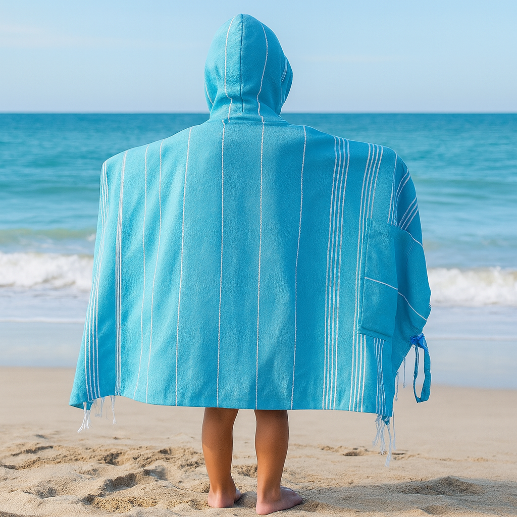 Child standing on a sandy beach wearing a Towlo blue 3-in-1 hooded towel shown fully open from the back, with ocean waves and a bright blue sky in the background