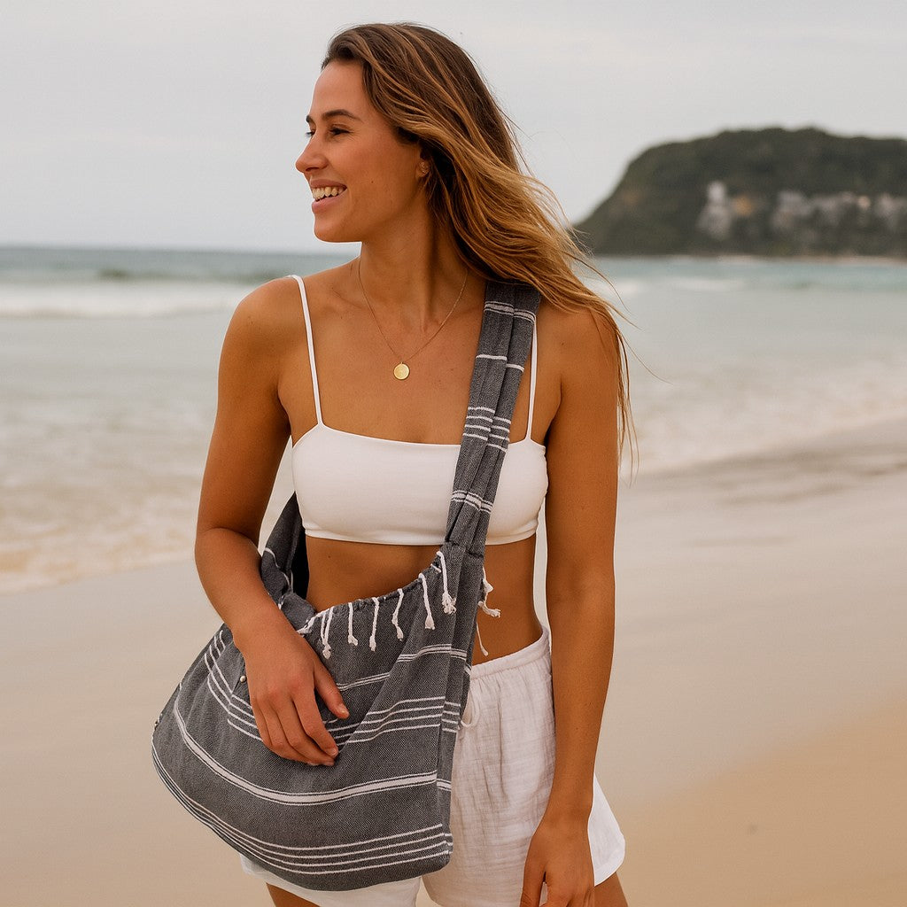 Woman smiling on a Gold Coast beach carrying a grey Towlo Turkish towel tote bag with white stripes over her shoulder.