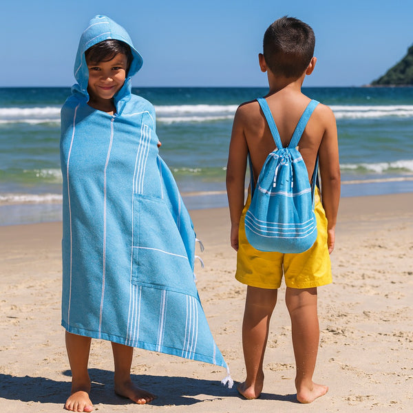 Two children on a beach wearing Towlo blue 3-in-1 hooded towel and matching towel backpack, shown from front and back with ocean waves in the background